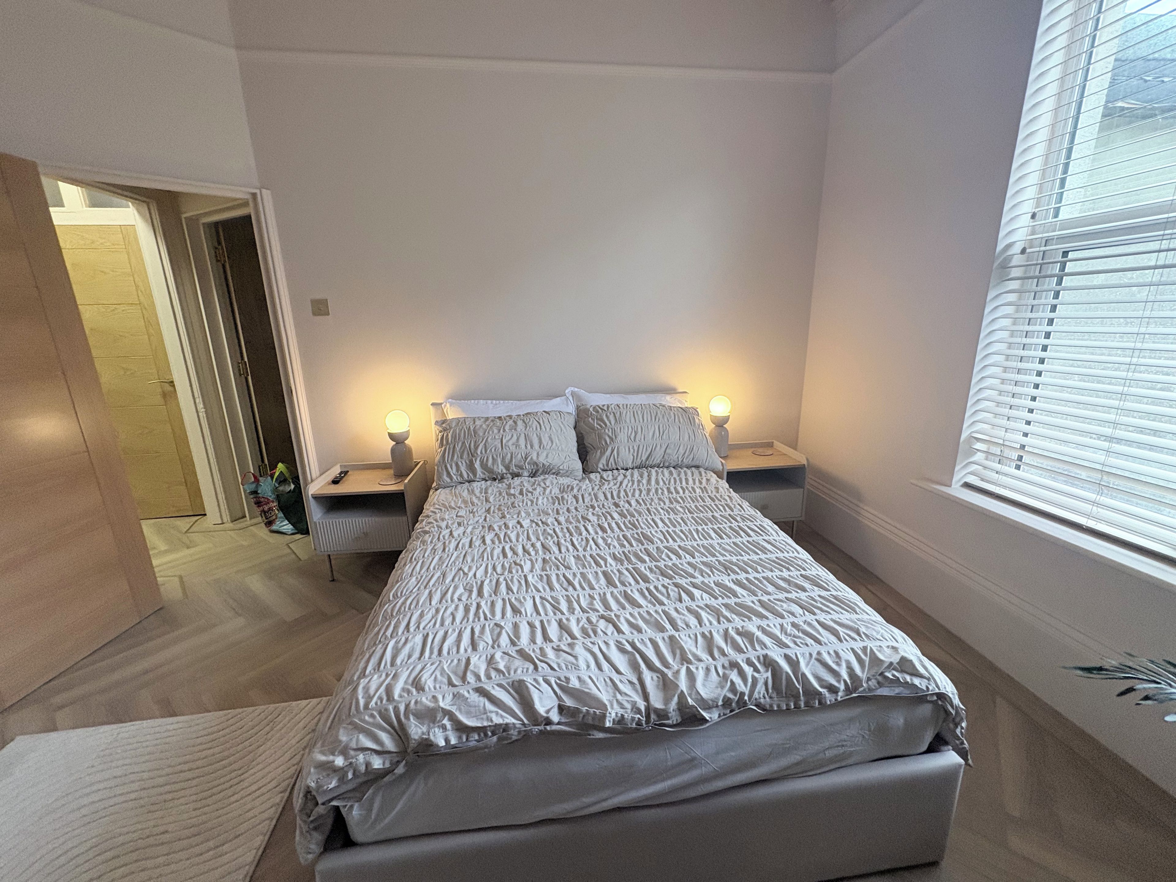 Une chambre à coucher comprenant un lit, deux tables de chevet, une commode, une porte et une fenêtre avec des stores.