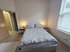 Une chambre à coucher comprenant un lit, deux tables de chevet, une commode, une porte et une fenêtre avec des stores.