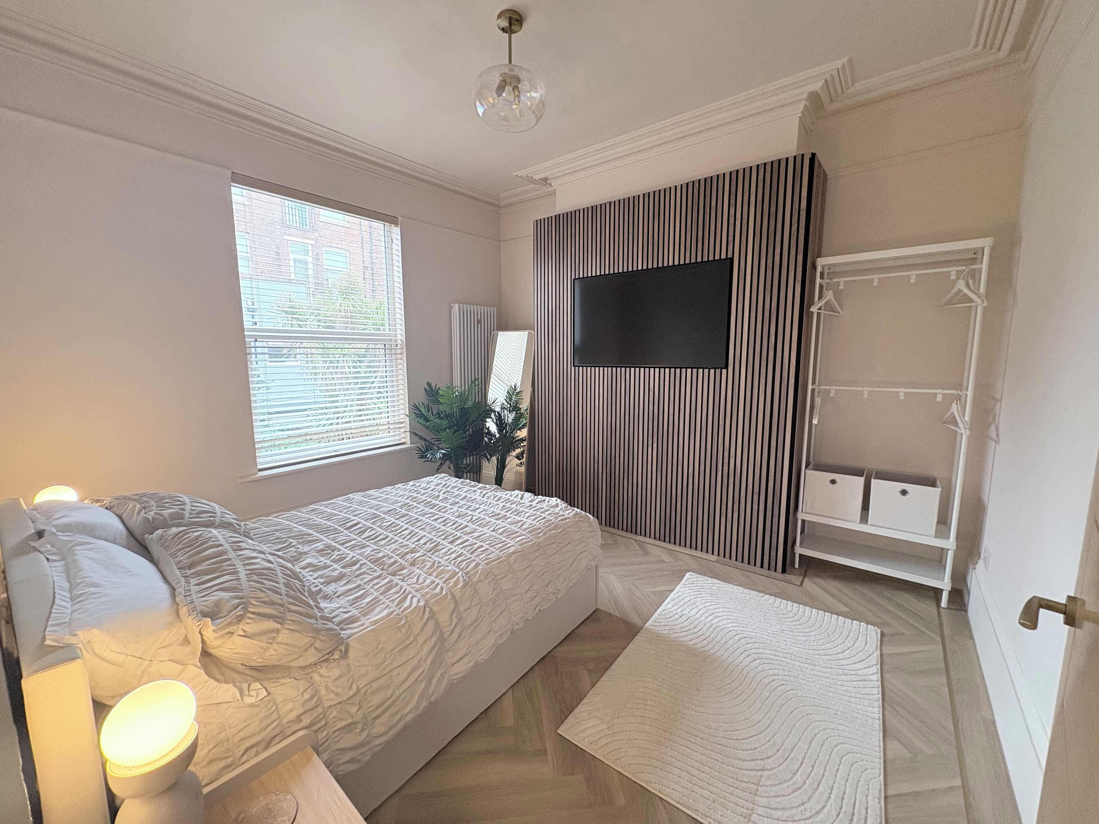 Un dormitorio con cama, lámpara de noche, ventana y televisión instalada en la pared.