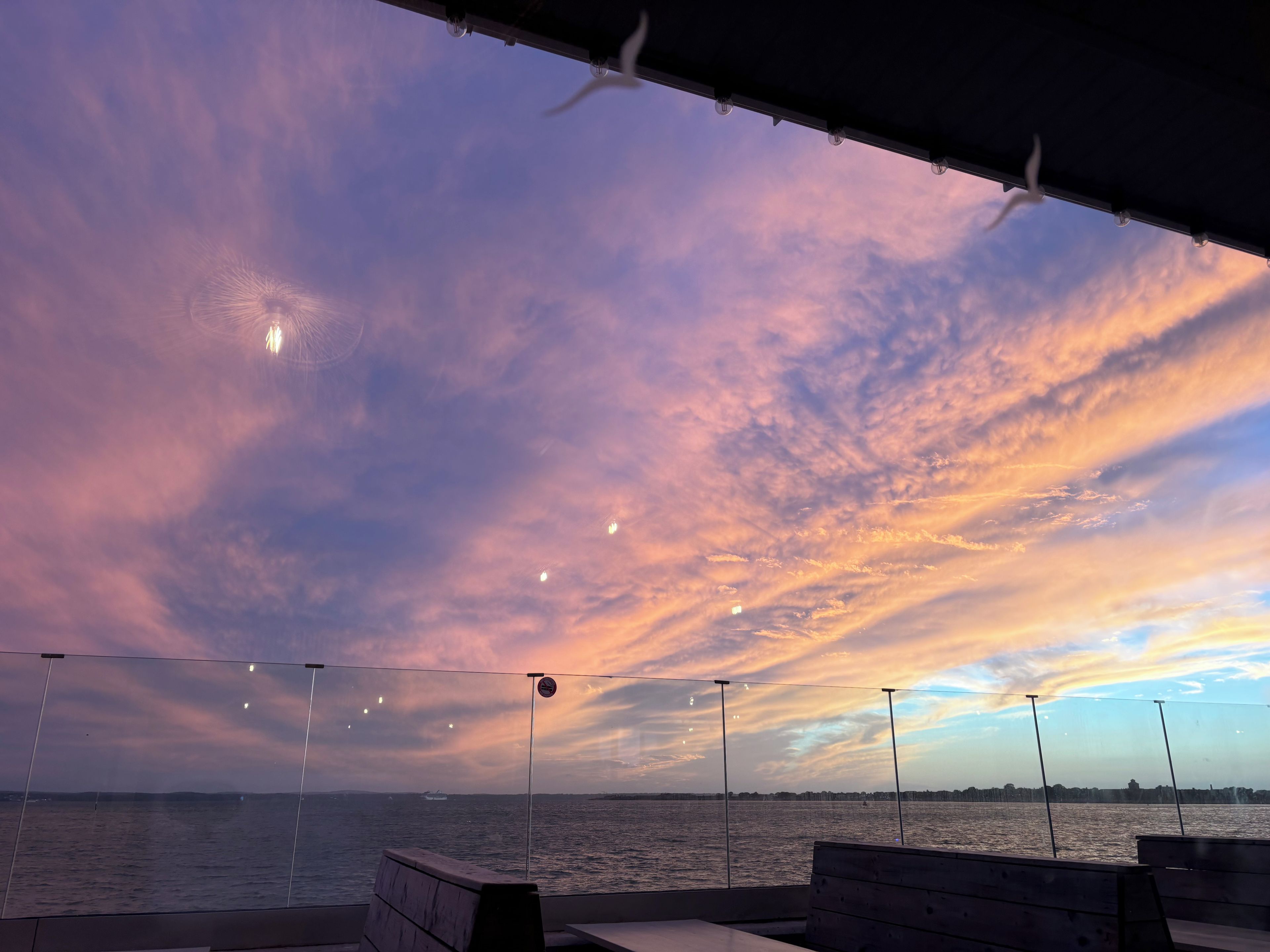 A view from a covered outdoor area with a bench, overlooking a body of water and a distant shoreline.
