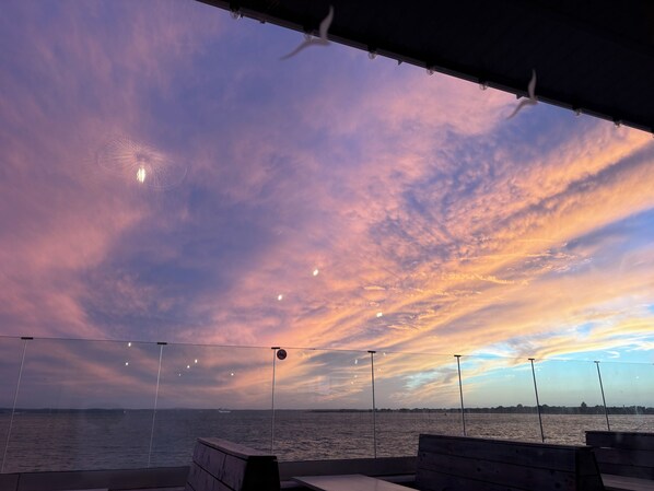 A view from a covered outdoor area with a bench, overlooking a body of water and a distant shoreline.