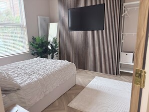Un dormitorio con una cama, televisor montado en la pared, un radiador y una planta.