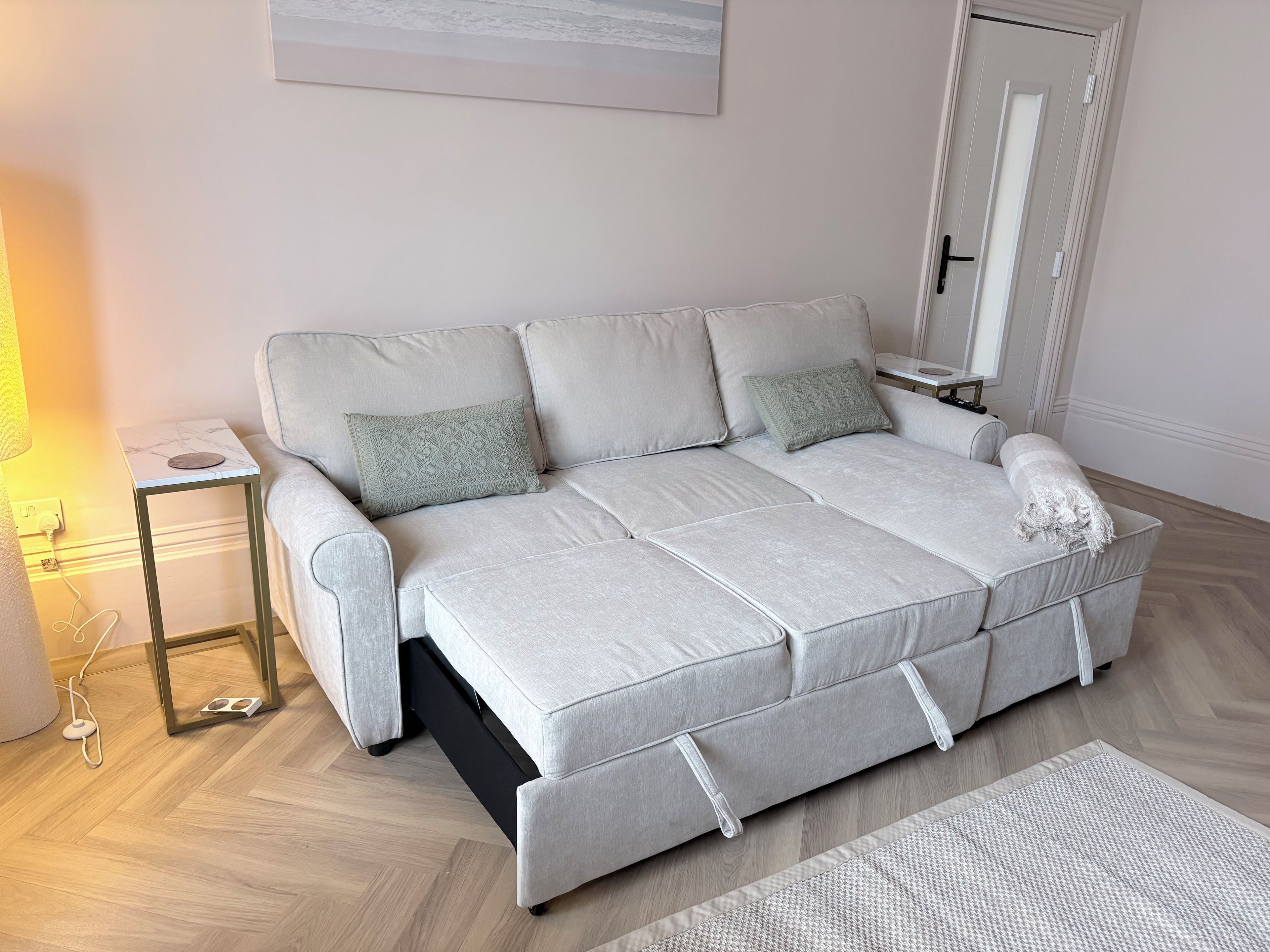 A light grey sofa with a matching throw blanket, a small side table with a lamp, and a woven rug on a wooden floor.