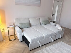 Un canapé gris clair, accompagné d’un plaid assorti, une petite table d’appoint avec une lampe et un tapis tissé posé sur un plancher en bois.