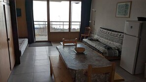 Ein Zimmer mit Esstisch, Stühlen, einer Couch, einem Kühlschrank und einem Fenster mit Vorhängen.