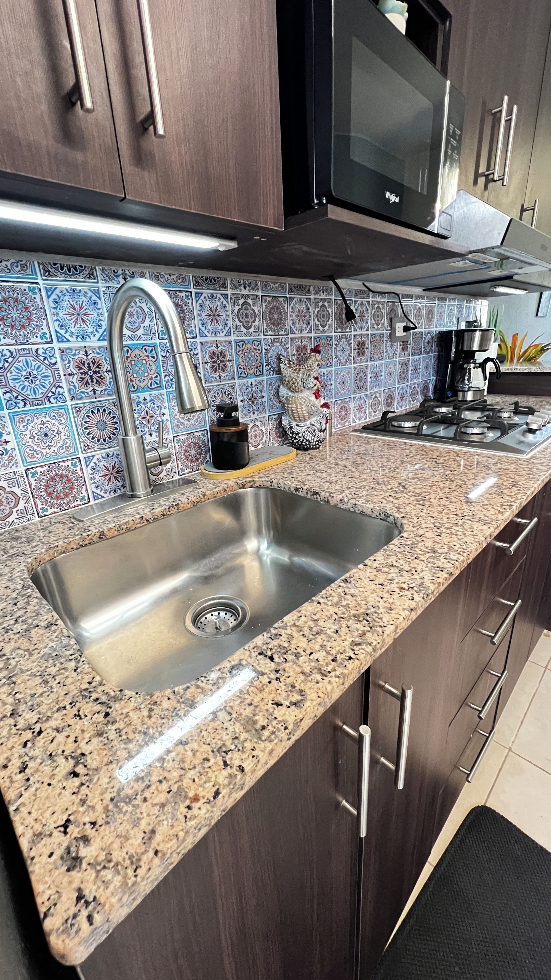 A kitchen with a stainless steel sink, dark wooden cabinets, a microwave, and a tiled backsplash.