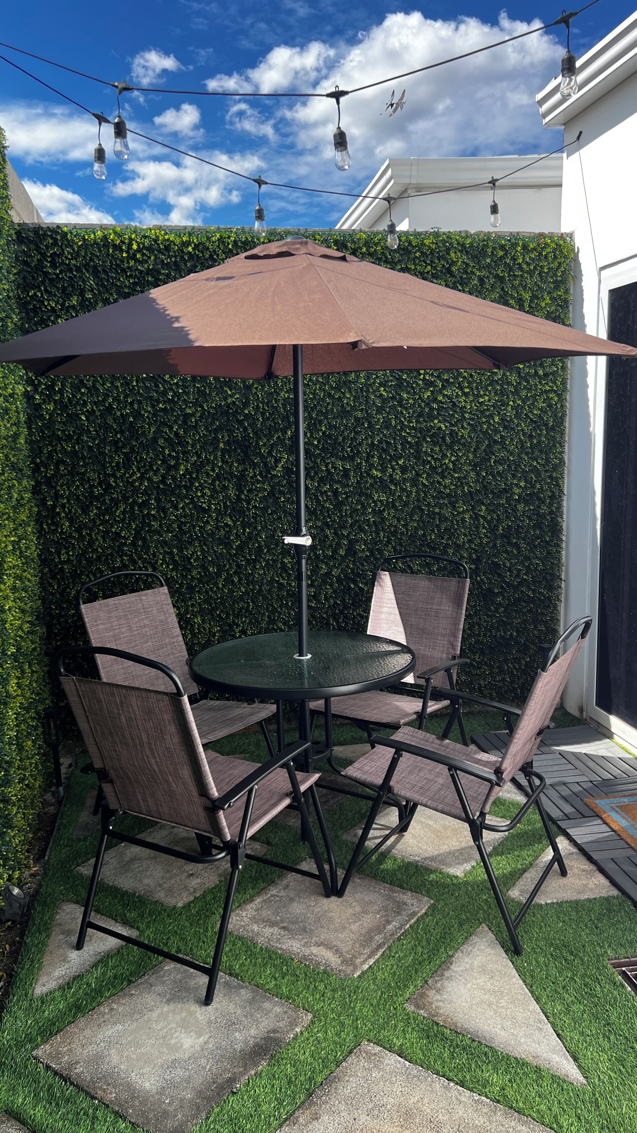 En terrasse med et rundt bord og stole, en brun parasol og en hæk.