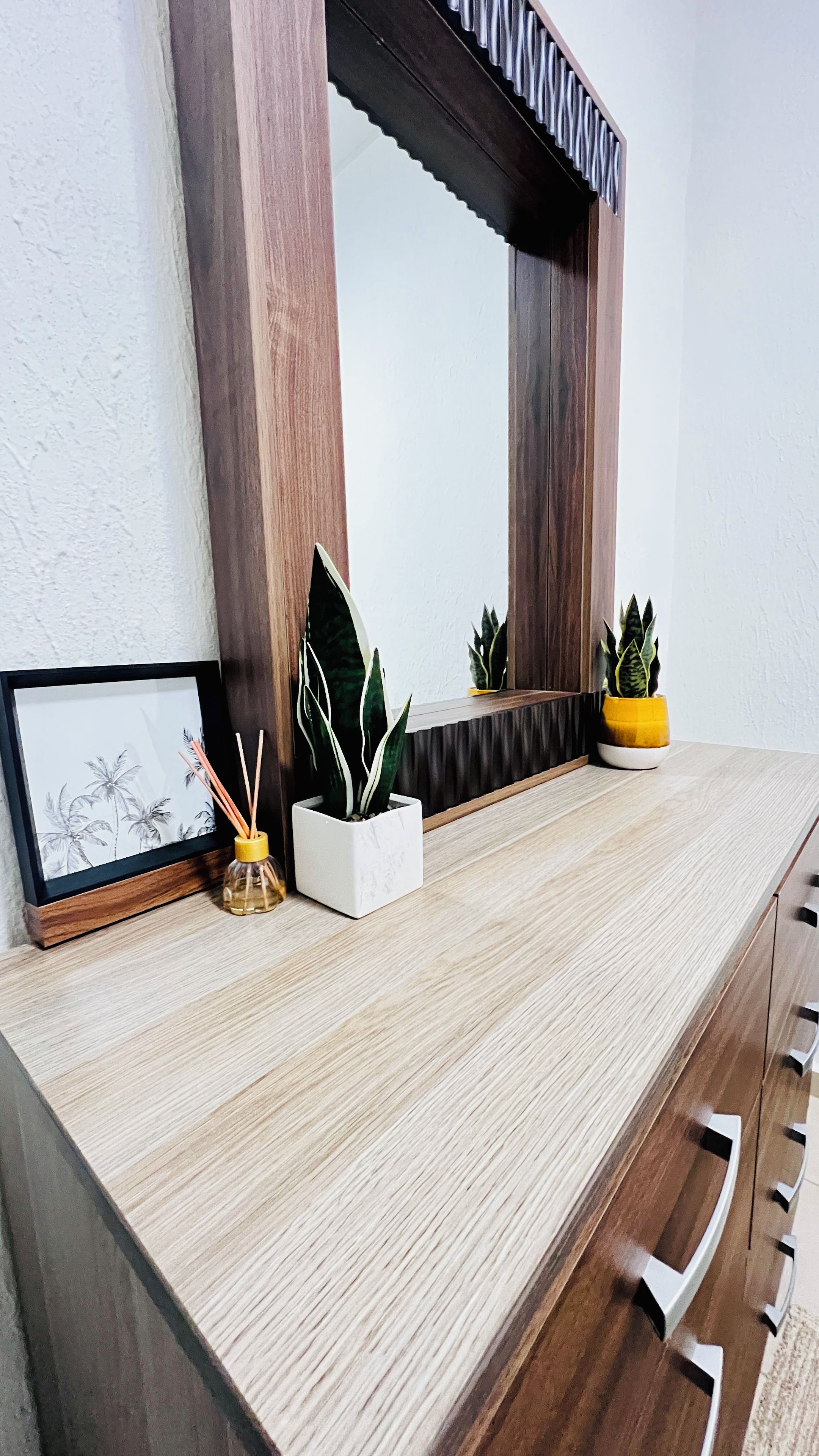 Une coiffeuse en bois avec un miroir, un tableau encadré et deux plantes en pot.
