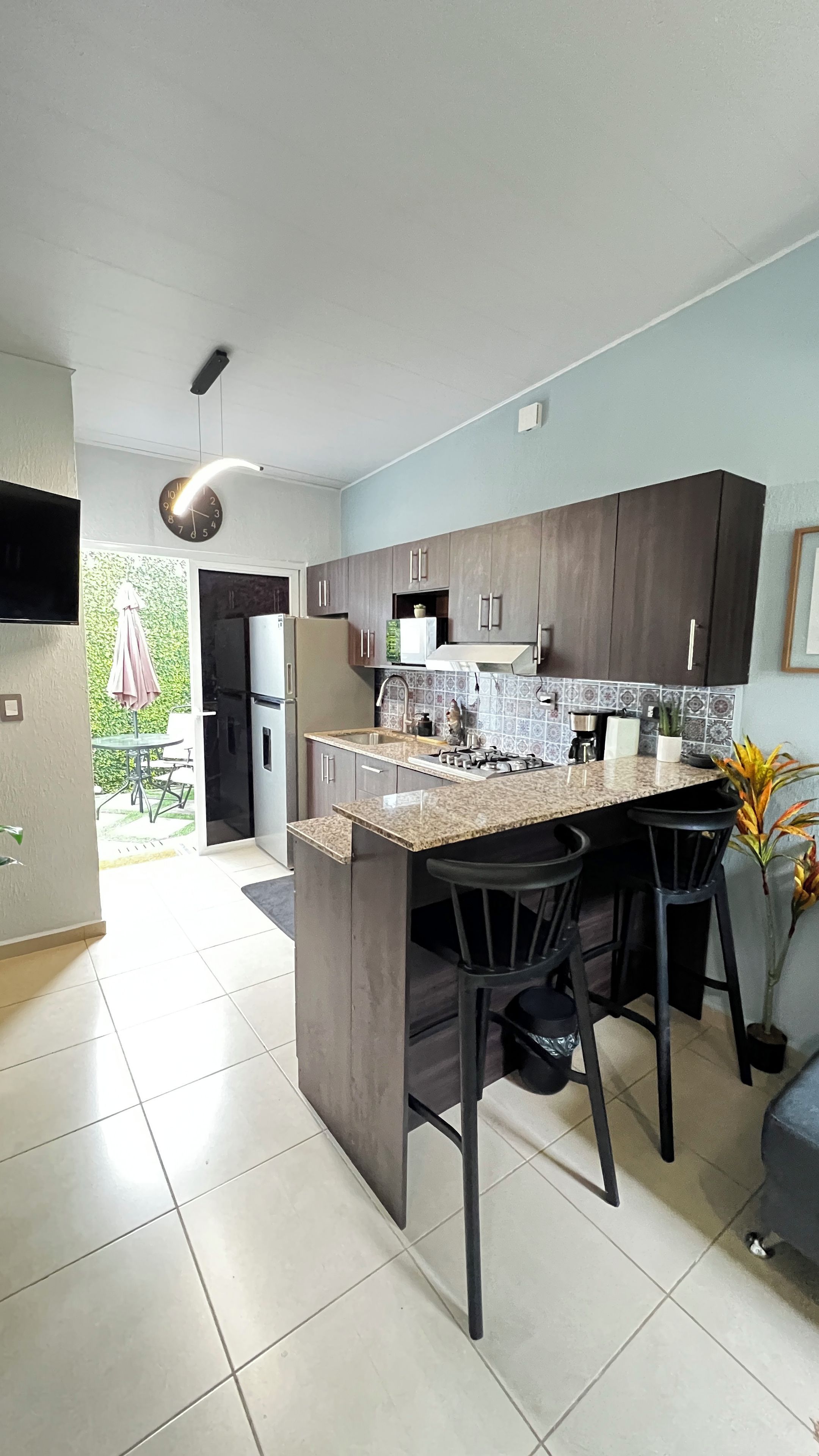 A modern kitchen with dark cabinets, a central island with bar stools, a built-in oven, and a visible clock.