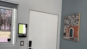 A room with a white door, a window with a view of trees, a mounted television, and a painting on the wall. - Contemporary Oasis, Serene Modern Retreat 2BR house with AC in vibrant Santa Ana (Santa Ana)