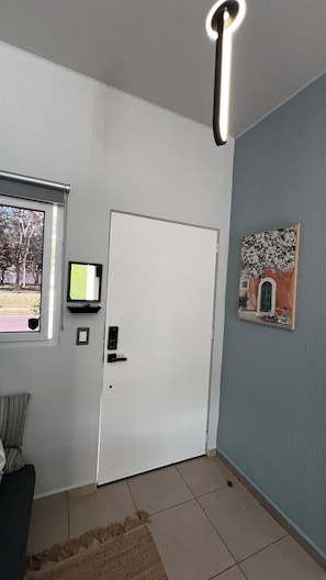 Een kamer met een witte deur, een raam met uitzicht op bomen, een wandtelevisie en een schilderij aan de muur.