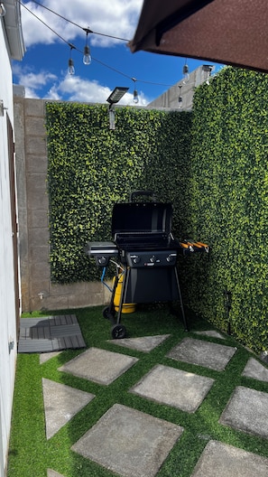 Een achtertuin met een barbecue, kunstgras, stenen looppaden en een haag.