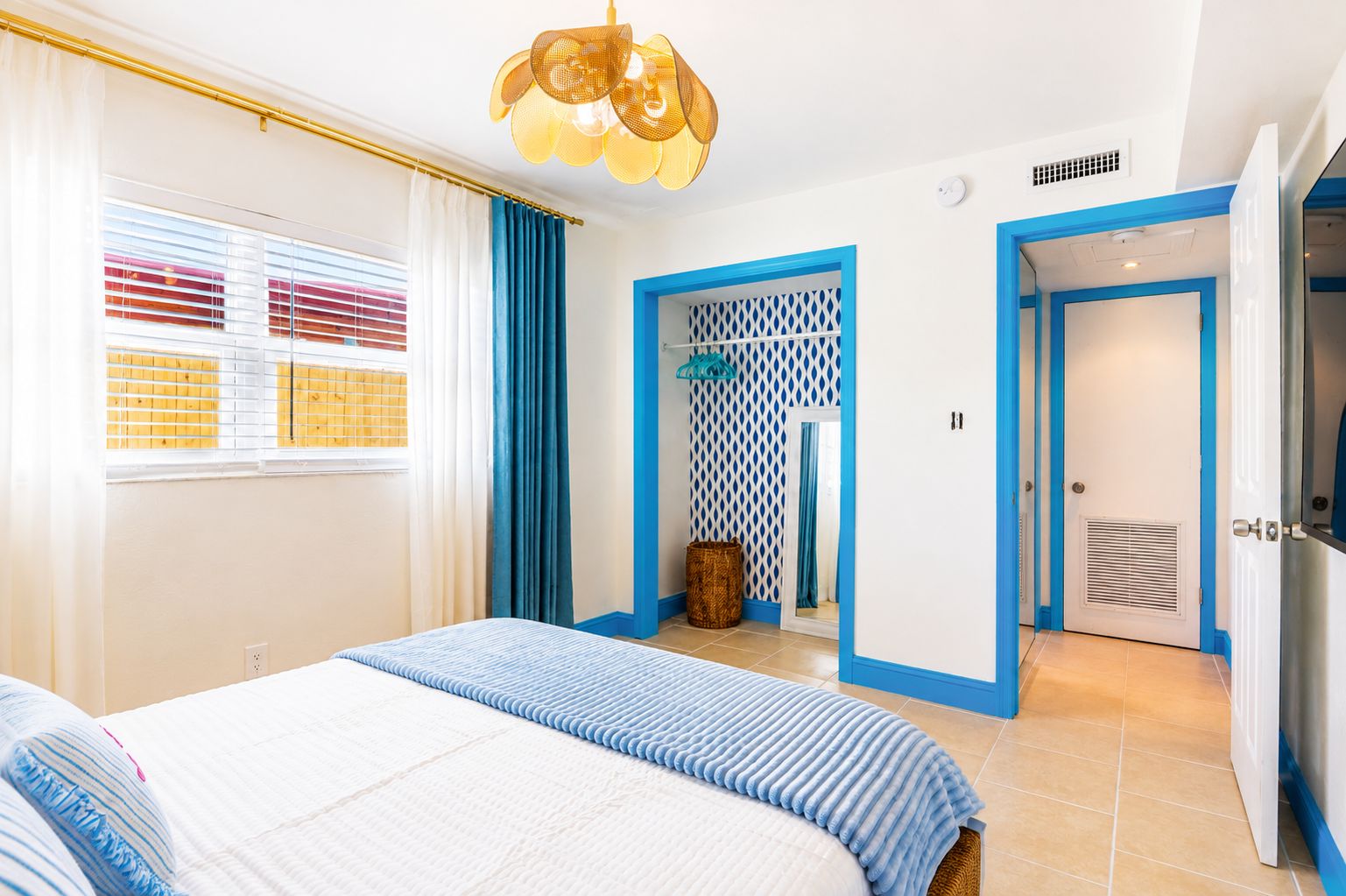 Une chambre d’hôtel avec un lit, une fenêtre avec des stores, une porte avec des moulures bleues et un luminaire au plafond.