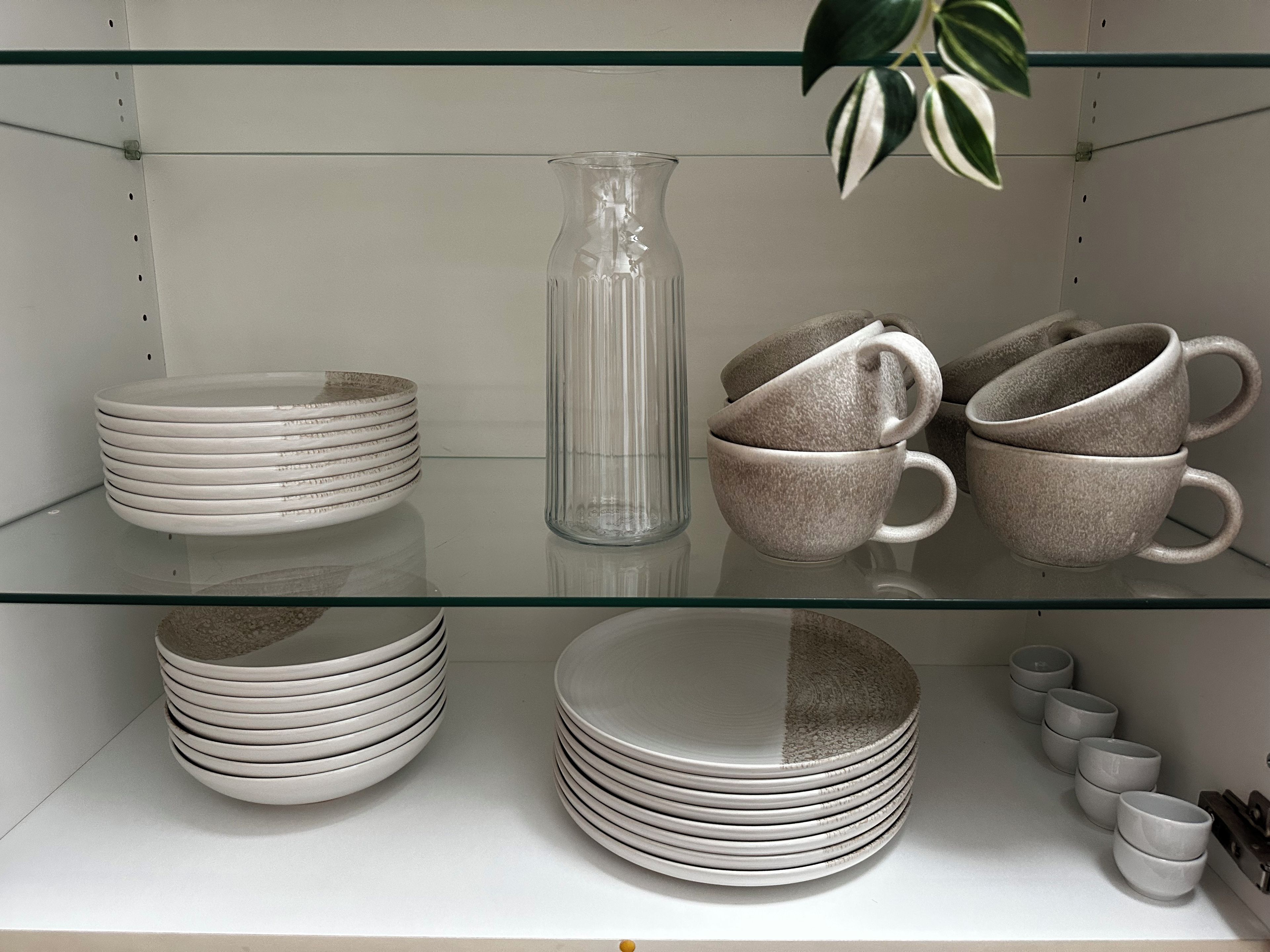 A glass shelf with stacked plates, a clear glass pitcher, and ceramic mugs.