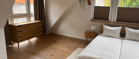 A bedroom with a wooden bed, a dresser, a nightstand, and a hanging plant.