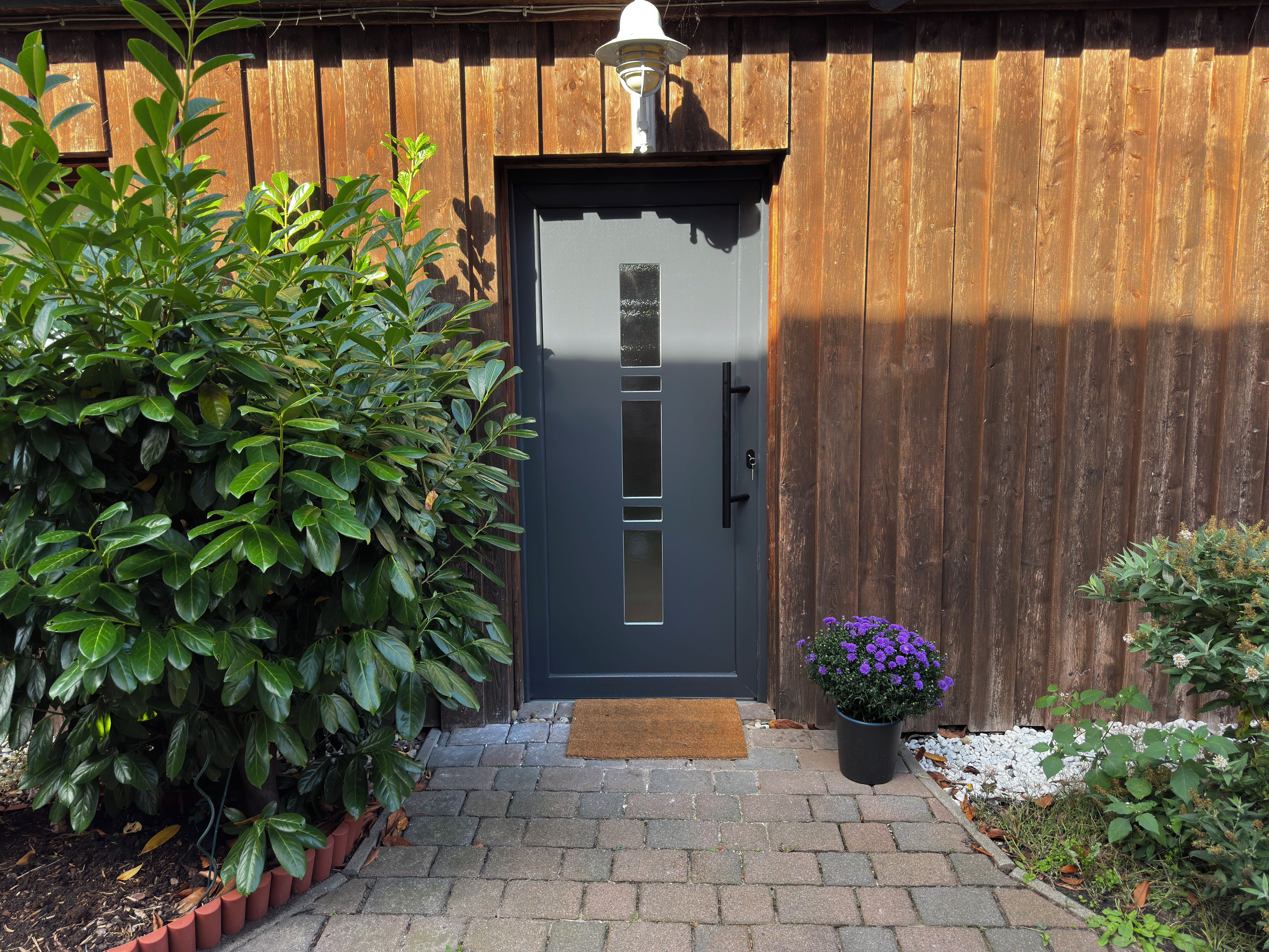 A wooden door with a dark-colored doorbell, a potted plant with purple flowers, and a doormat on a brick pathway.