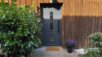 A wooden door with a dark-colored doorbell, a potted plant with purple flowers, and a doormat on a brick pathway.