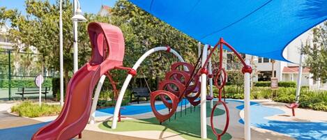 A playground with a slide and climbing structures, surrounded by greenery and a blue canopy.