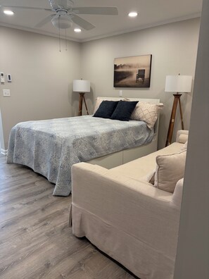 A bedroom with a bed, a nightstand, a floor lamp, a ceiling fan, and a painting on the wall. - Beautiful 1-bedroom condo in beach town Wells with an indoor pool (Wells)