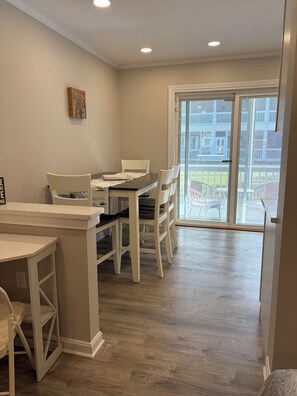 A dining area with a table and chairs, a white wall, and a view through a glass door to an outdoor deck. - Beautiful 1-bedroom condo in beach town Wells with an indoor pool (Wells)