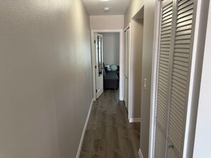 Un couloir doté d’un plancher en bois, de murs blancs et d’une porte donnant sur une chambre à coucher, où se trouvent un lit et une fenêtre.