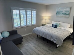 Une chambre à coucher avec un lit, une table de chevet, une lampe, une fenêtre avec des stores, un canapé et un tableau au mur.