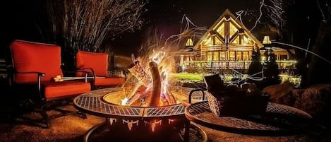 A fire pit with seating, a large house in the background, and a well-lit outdoor area.