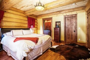 Ein Schlafzimmer in einer Blockhütte mit einem großen Bett, Holzwänden, einer Deckenleuchte, einem Schminktisch und einer Tür.