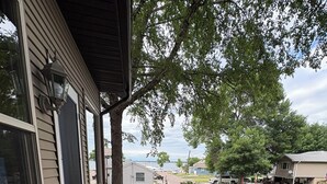 Une terrasse en bois avec un banc, une maison aux murs recouverts d’un revêtement beige, et une étendue d’eau visible au loin.