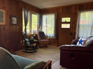 Ein gemütliches Wohnzimmer mit Holzwänden, einem Schaukelstuhl, einem karierten Sessel, einem Ledersofa mit einem Sternenkissen und einer Tür mit Blick aus dem Fenster.