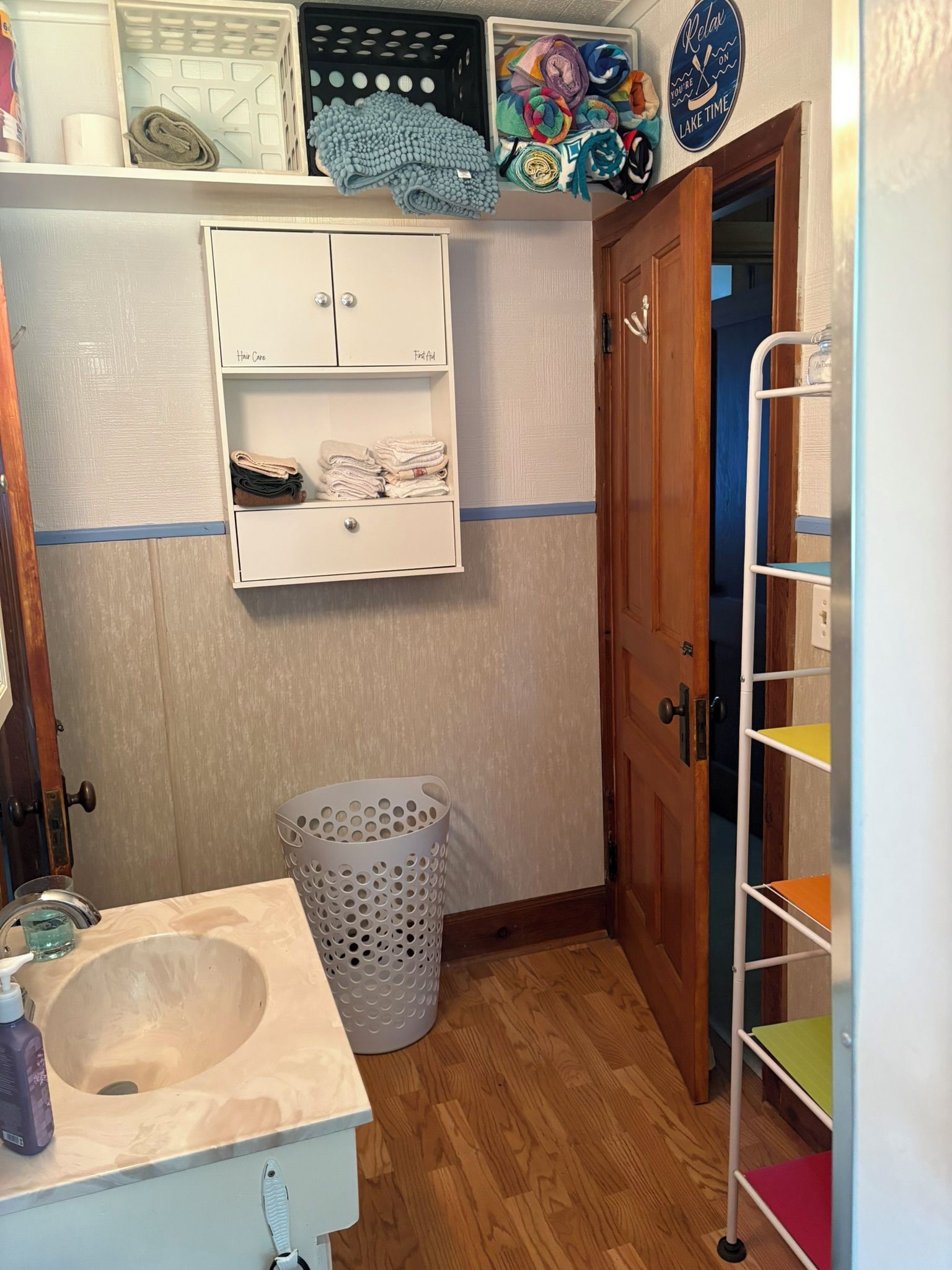 Une salle de bain avec un lavabo, un panier à linge, une étagère avec des serviettes pliées et une porte donnant sur une autre pièce.