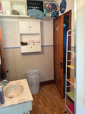 Un bany amb un rentaplat, una cistella de roba, una prestatgeria amb tovalloles doblegades i una porta que dona a una altra habitació.