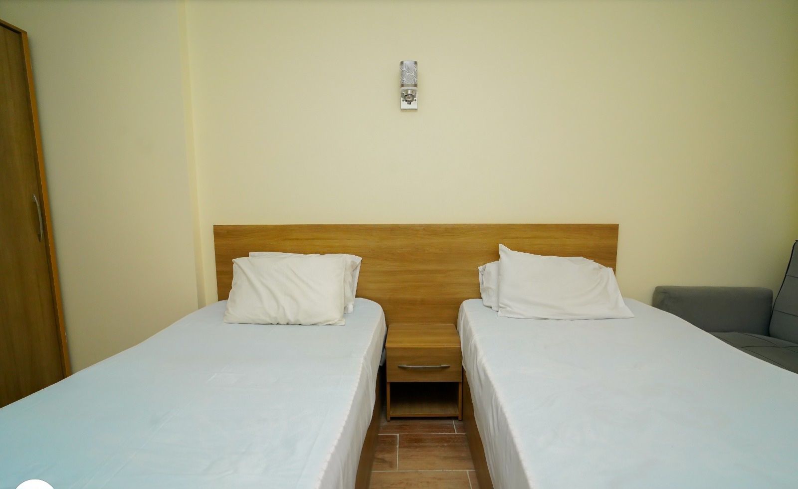 A hotel room with two single beds, a wooden headboard, a small bedside table, and a grey sofa.
