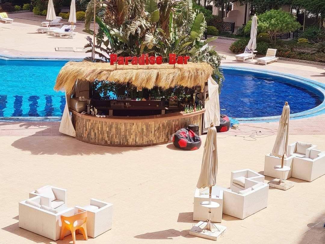 Un espace bar aménagé au bord de la piscine, doté d’un toit de chaume, de chaises longues blanches et de parasols.