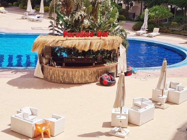 Un espace bar aménagé au bord de la piscine, doté d’un toit de chaume, de chaises longues blanches et de parasols.