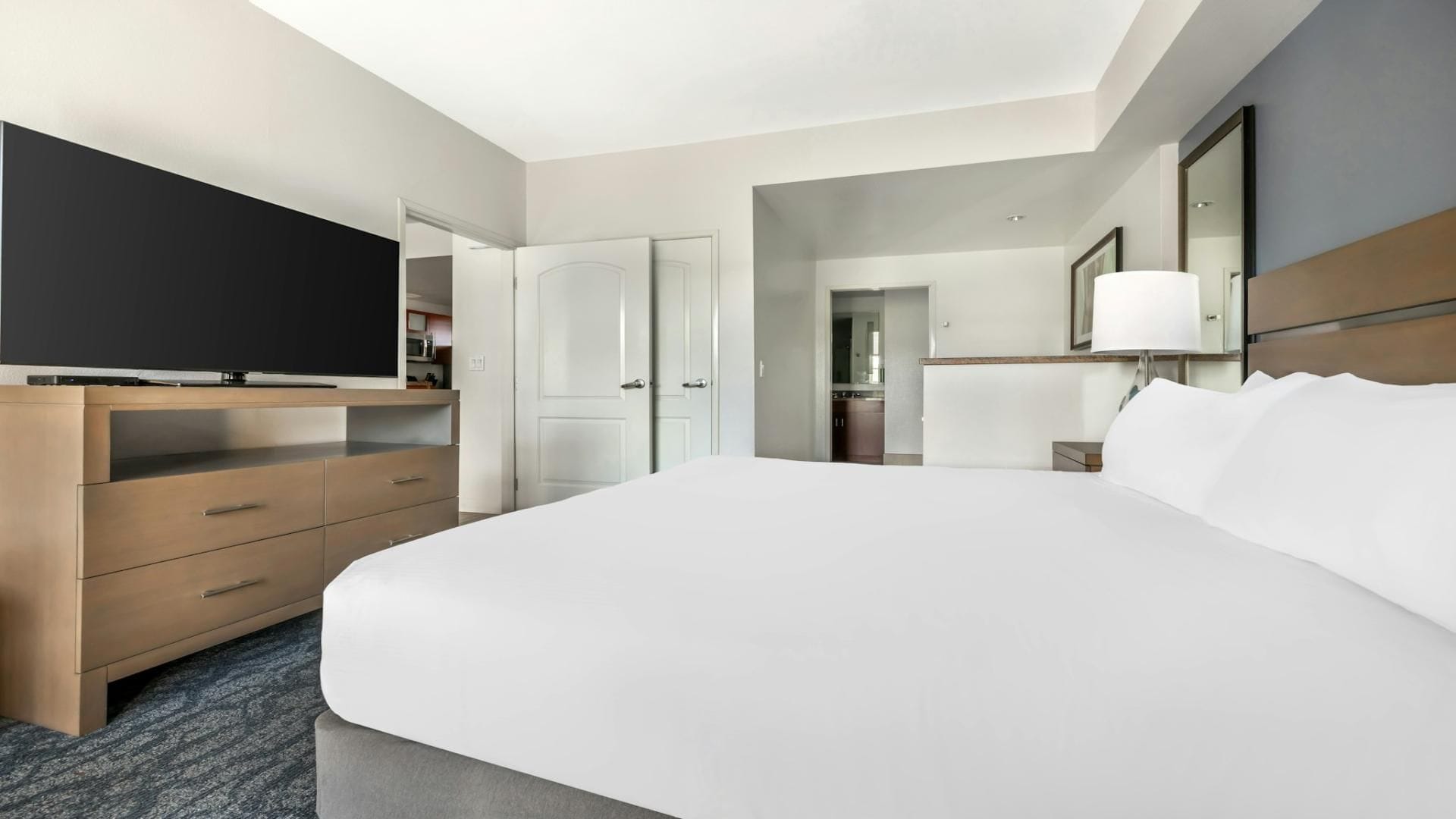 A hotel room with a large bed, a television on a wooden dresser, a desk with a lamp, and a door leading to a bathroom.