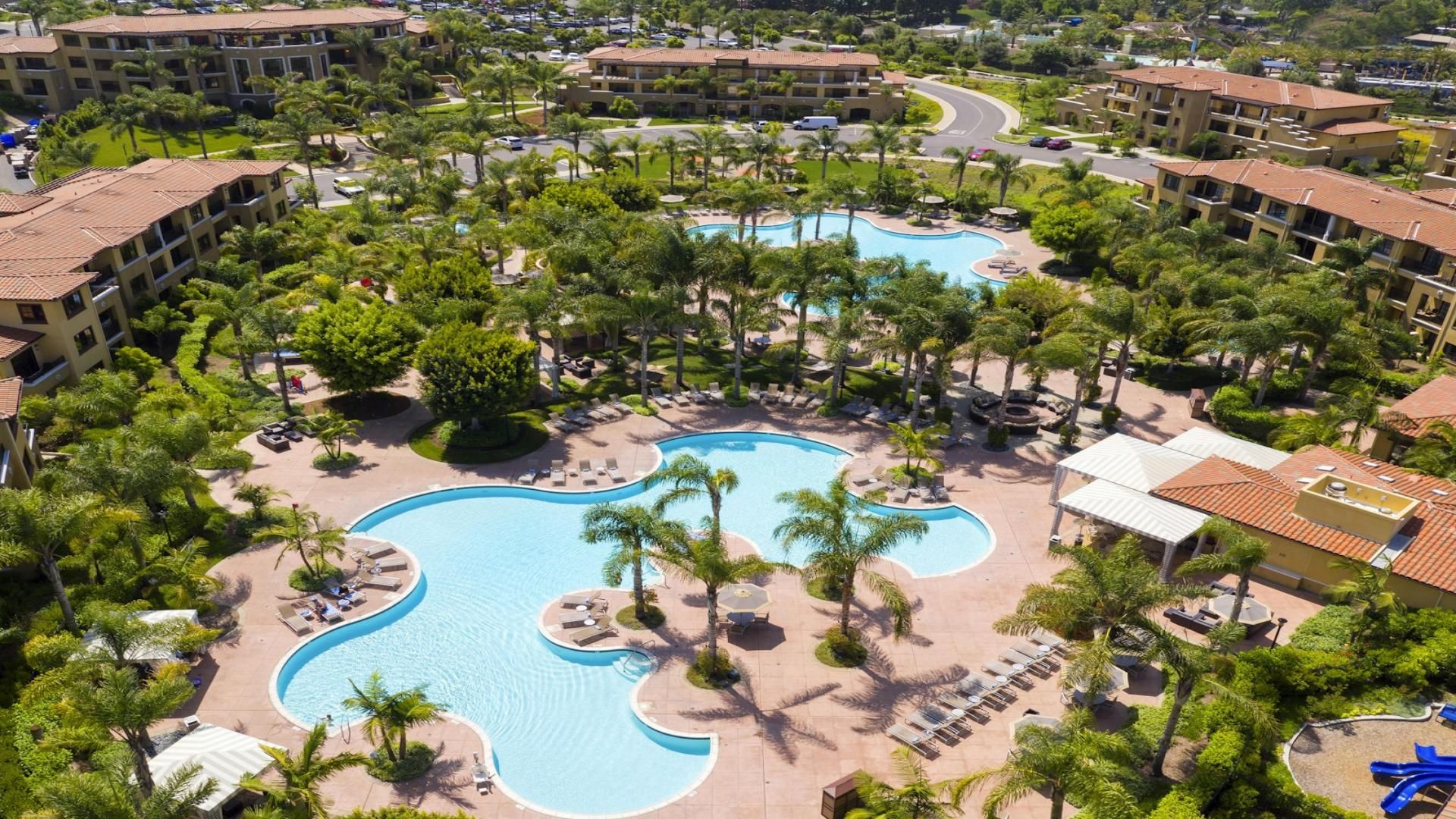 Un complexe hôtelier doté d’une piscine centrale, entourée de palmiers et agrémenté d’espaces de détente.