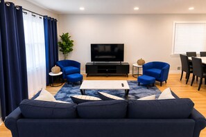 A modern living room with a sofa, two armchairs, a coffee table, a TV, and a dining area with chairs and a table. - Spacious 5BR, Near NYC, Reserved Parking & Yard (Jersey City)