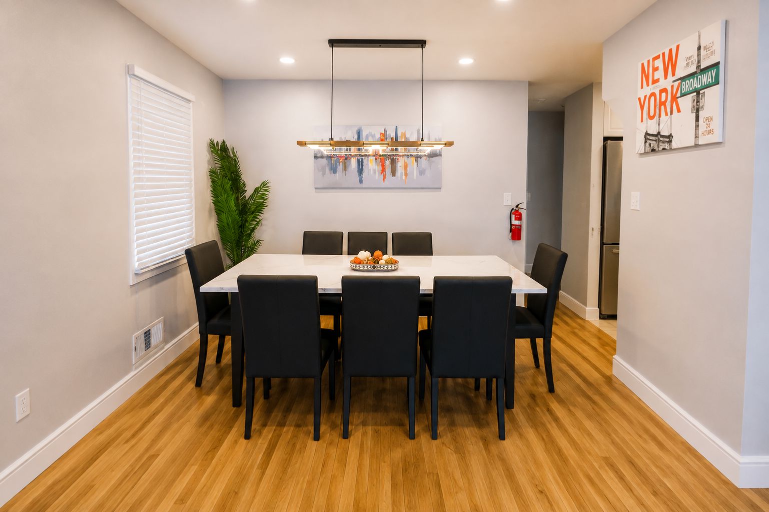 A dining area with a white table, black chairs, a modern pendant light, and a painting of a cityscape.