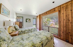 A bedroom with two beds, a wooden wall, a window with a view of greenery, and a small table with a lamp.