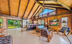 A spacious interior with a high ceiling, wooden walls, a dining area, and a view of greenery outside.