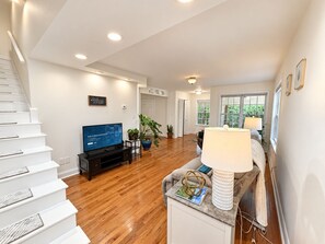 A spacious living area with a wooden staircase, a flat-screen TV, a coffee table, and a lamp.
