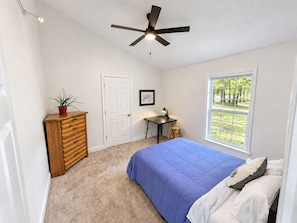 A bedroom with a bed, a desk, a chair, a ceiling fan, a window, and a door.