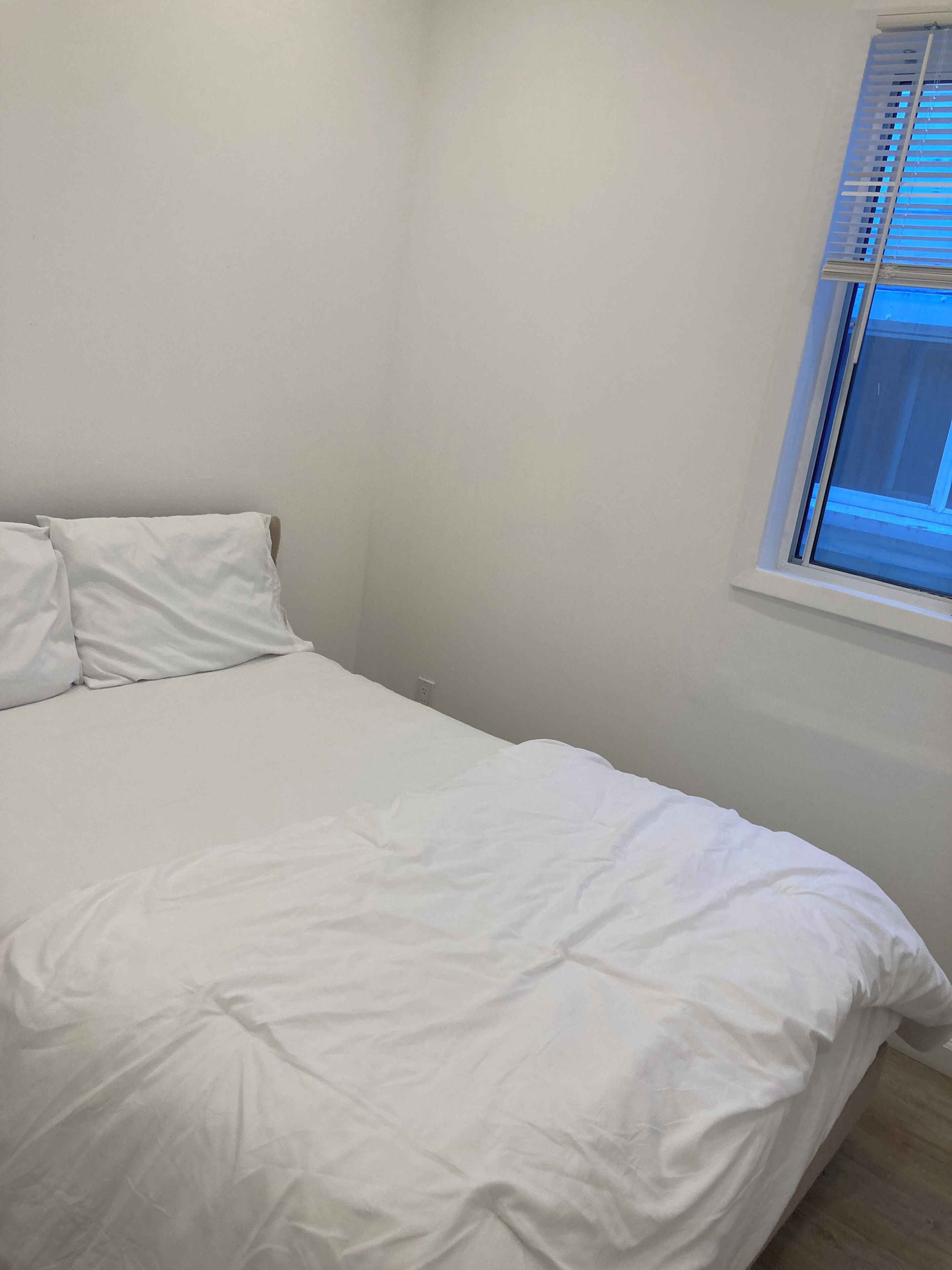 A single bed with white bedding in a room with white walls and a window with blinds.