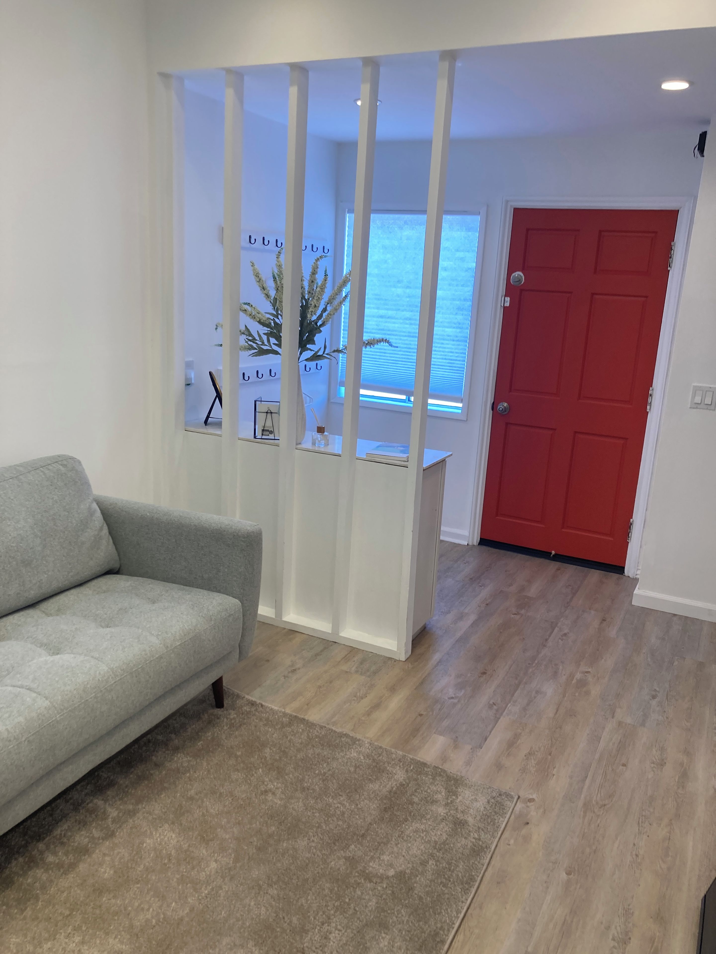 Una habitación con sofá gris, suelo de madera, puerta roja y ventana con vistas.