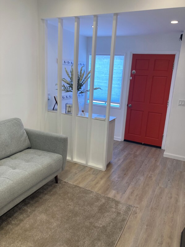Una habitación con sofá gris, suelo de madera, puerta roja y ventana con vistas.