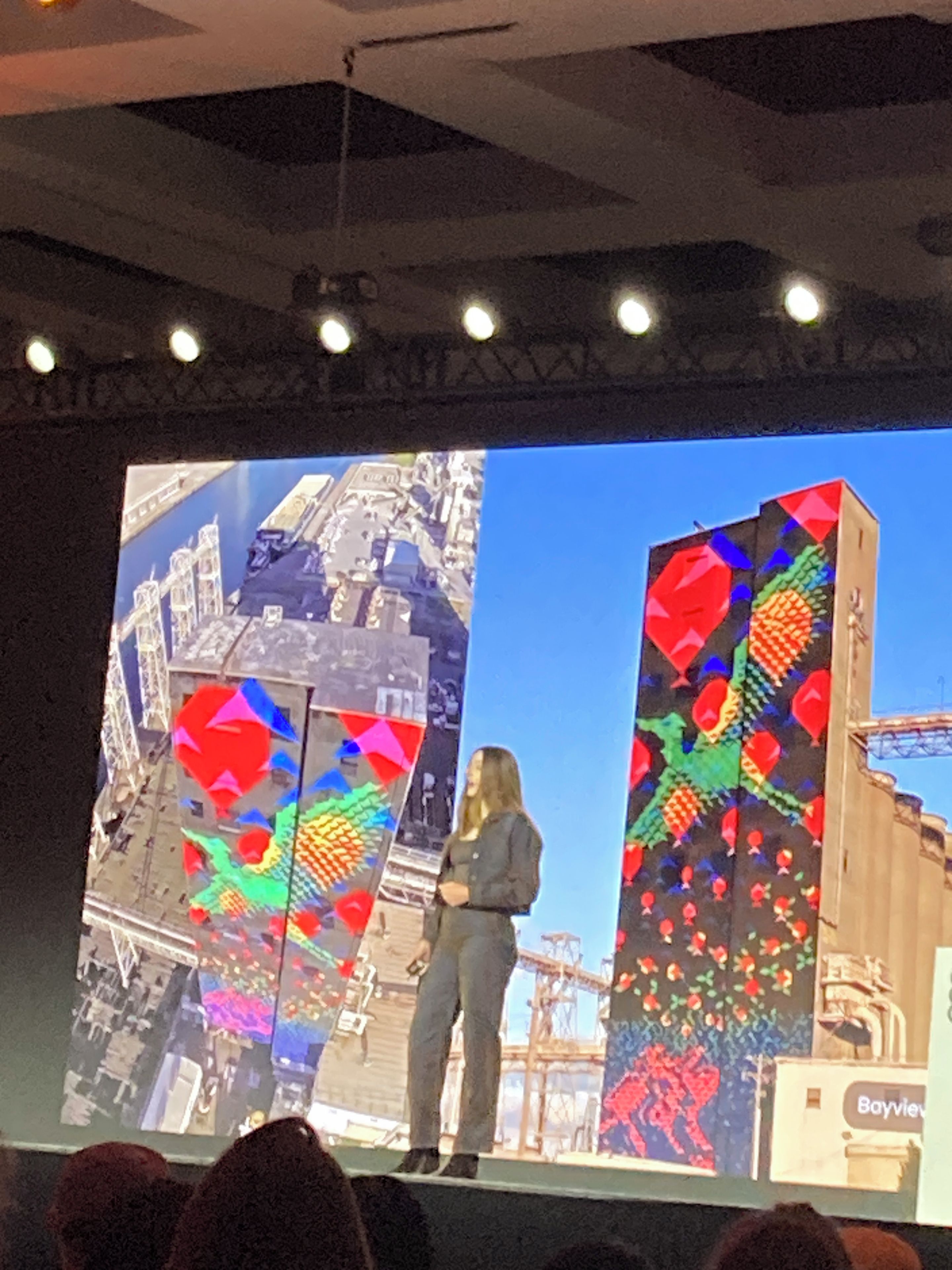 A person standing on a stage with a large screen displaying colorful geometric patterns.