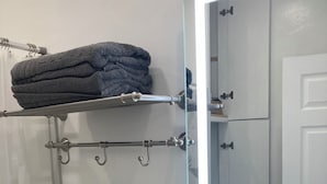 Une salle de bain comprenant des toilettes, un lavabo, un miroir, un porte-serviettes avec des serviettes pliées, une étagère avec une plante et une cabine de douche en verre.