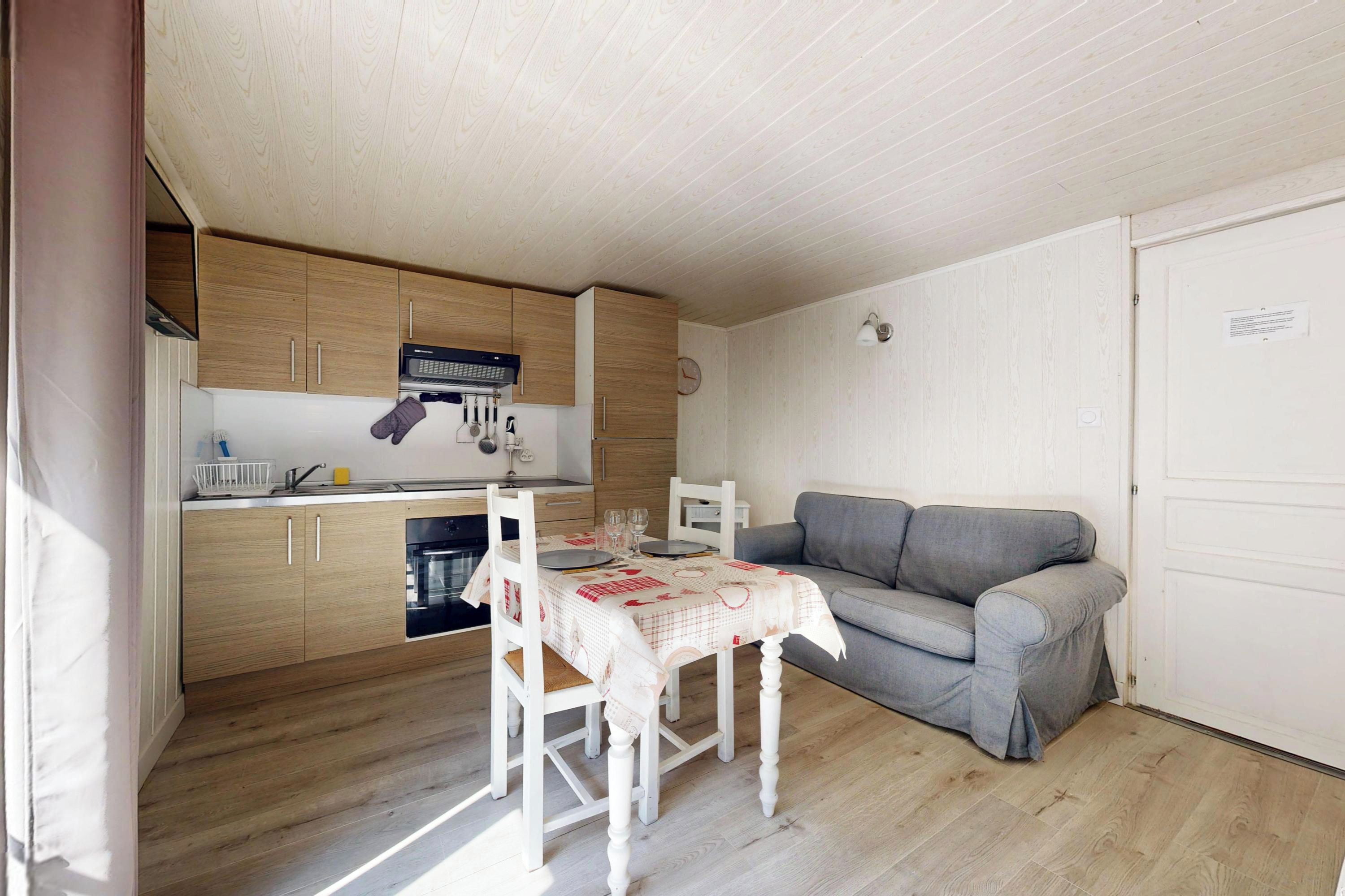 Een kleine keuken met houten keukenkastjes, een eettafel gedekt met borden en glazen, en een grijze bank in de aangrenzende woonkamer.