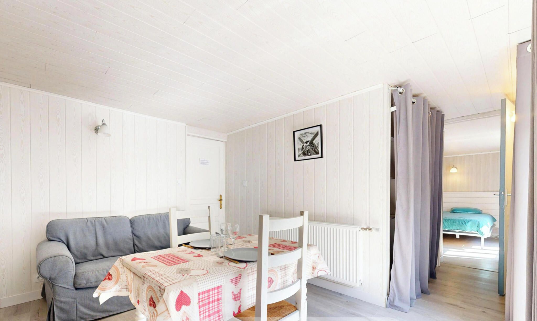 A small, cozy room with a grey sofa, a dining table set for two, and a bed in the background.